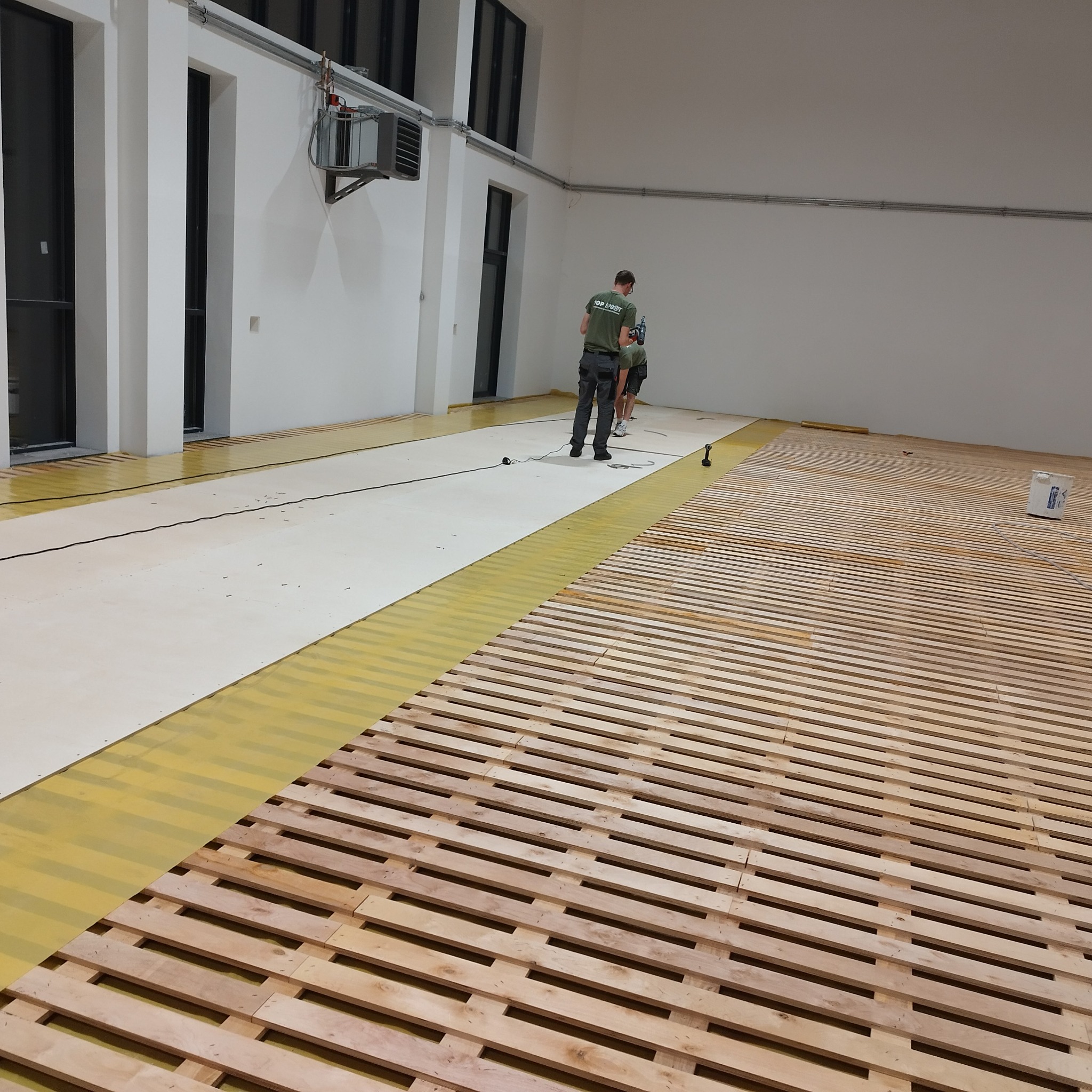 Montaż podłogi: drewniany ruszt pod podłogę w hali sportowej, widoczne narzędzia i ekipa montażowa w tle.