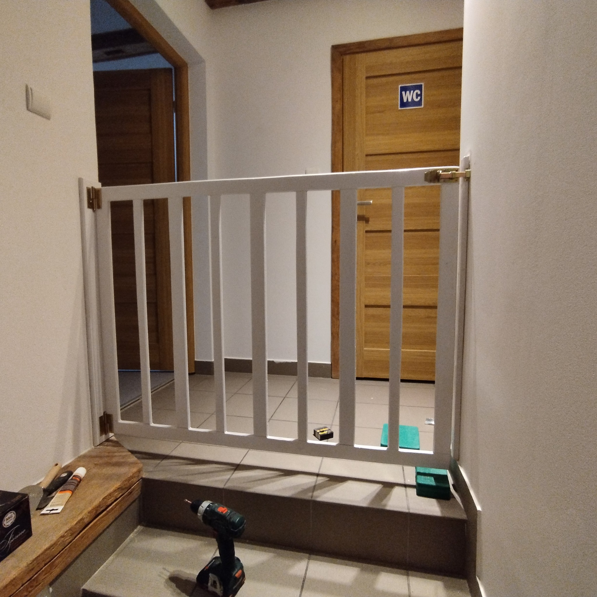 Biała barierka zabezpieczająca zamontowana na schodach, widoczna w tle drewniana ościeżnica z drzwiami oznaczonymi symbolem WC, w pobliżu narzędzia i akcesoria montażowe.