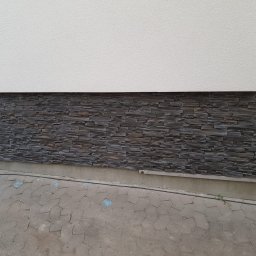 WIMIGA-BUD Łukasz Waniowski - Fragment elewacji budynku z dekoracyjnym kamieniem elewacyjnym w odcieniach szarości, ułożonym na dolnej części ściany, powyżej biały tynk strukturalny, widoczny fragment chodnika z szarej kostki...