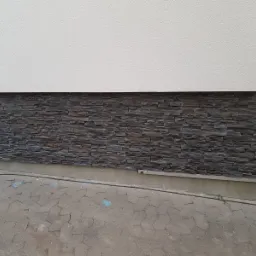 Fragment elewacji budynku z dekoracyjnym kamieniem elewacyjnym w odcieniach szarości, ułożonym na dolnej części ściany, powyżej biały tynk strukturalny, widoczny fragment chodnika z szarej kostki...