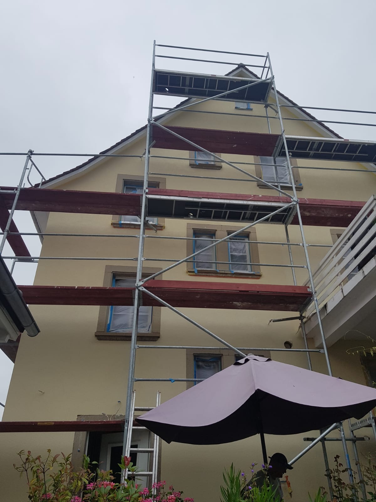 Dwupiętrowy dom w trakcie prac elewacyjnych, okna zabezpieczone folią, ustawione rusztowanie, widoczny fragment parasola ogrodowego i roślinność.