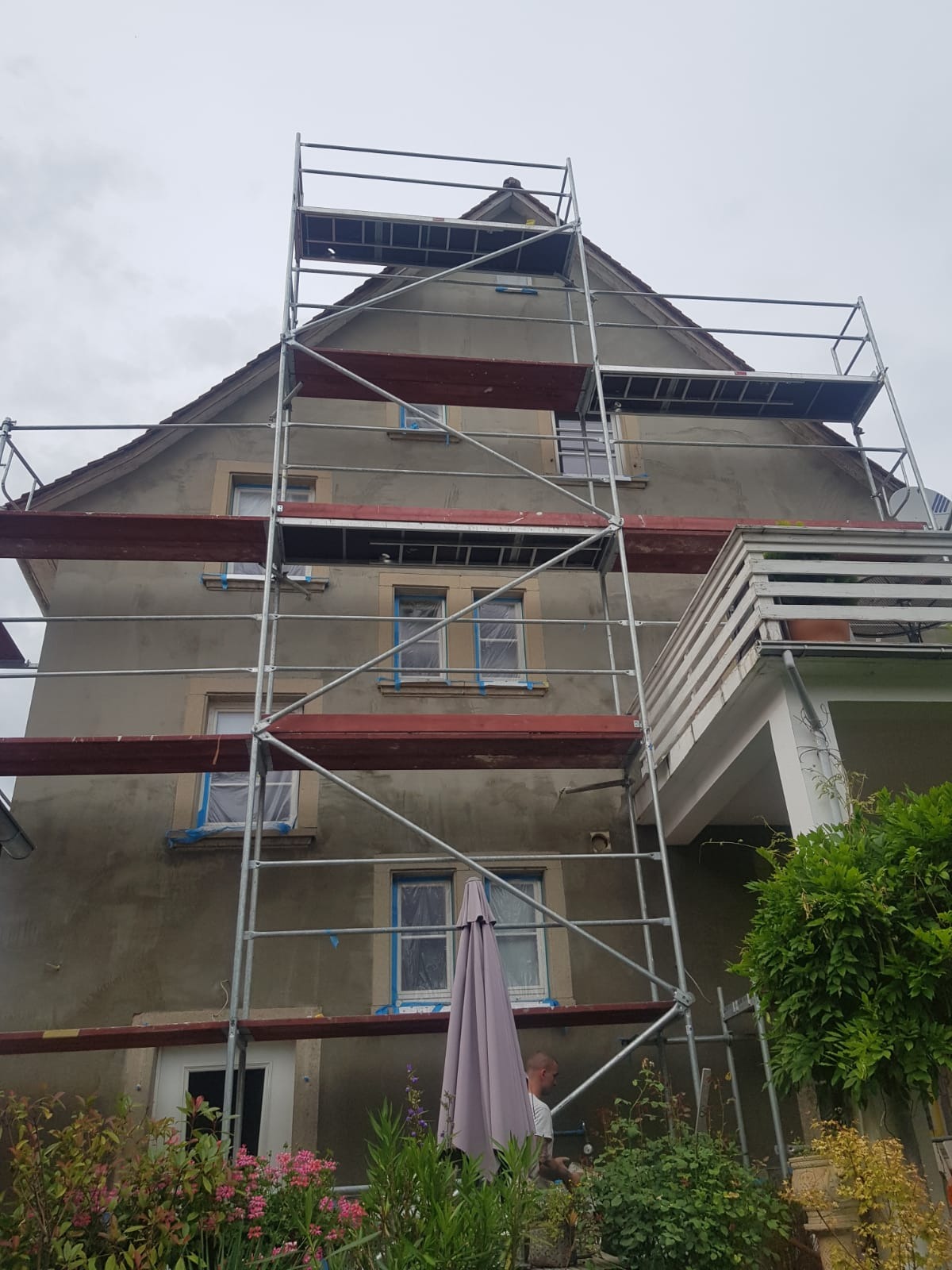 Dwupiętrowy dom w trakcie renowacji elewacji, z ustawionym rusztowaniem, okna zabezpieczone folią, widoczne rośliny w ogrodzie i fragment parasola.