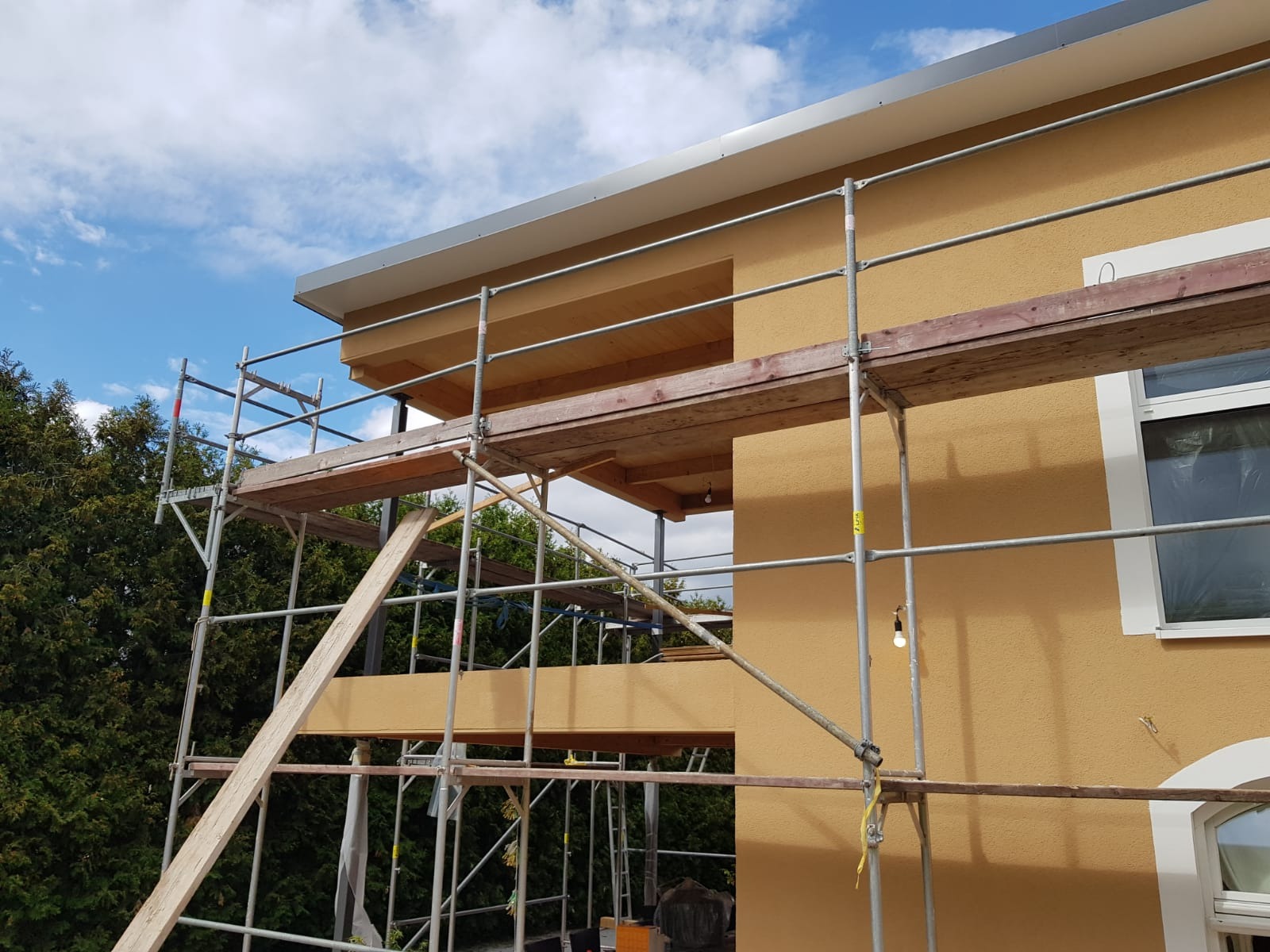 Ocieplona elewacja budynku w trakcie prac wykończeniowych, widoczne rusztowania i fragment okna z folią ochronną.