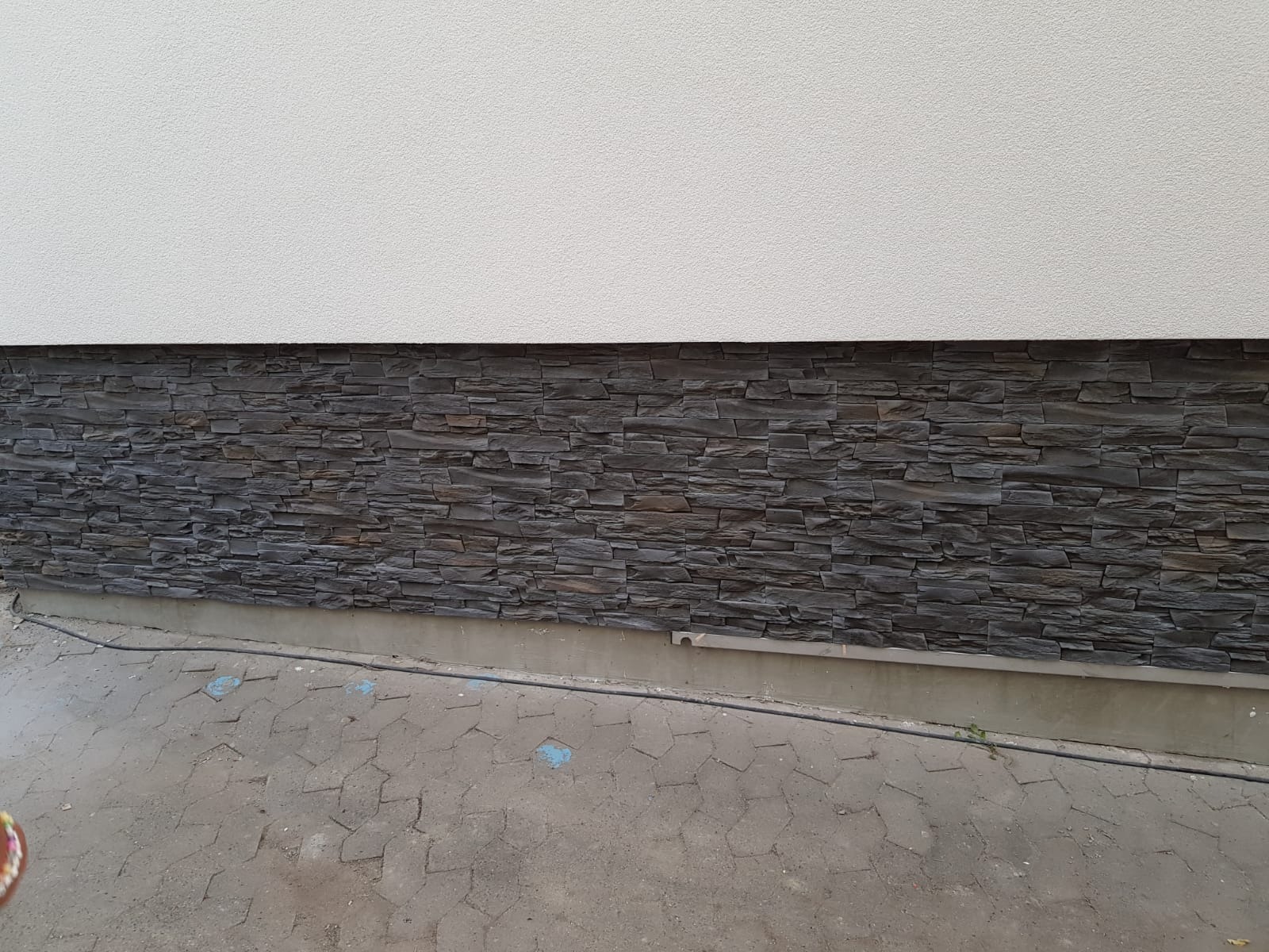 Fragment elewacji budynku z dekoracyjnym kamieniem elewacyjnym w odcieniach szarości, ułożonym na dolnej części ściany, powyżej biały tynk strukturalny, widoczny fragment chodnika z szarej kostki...