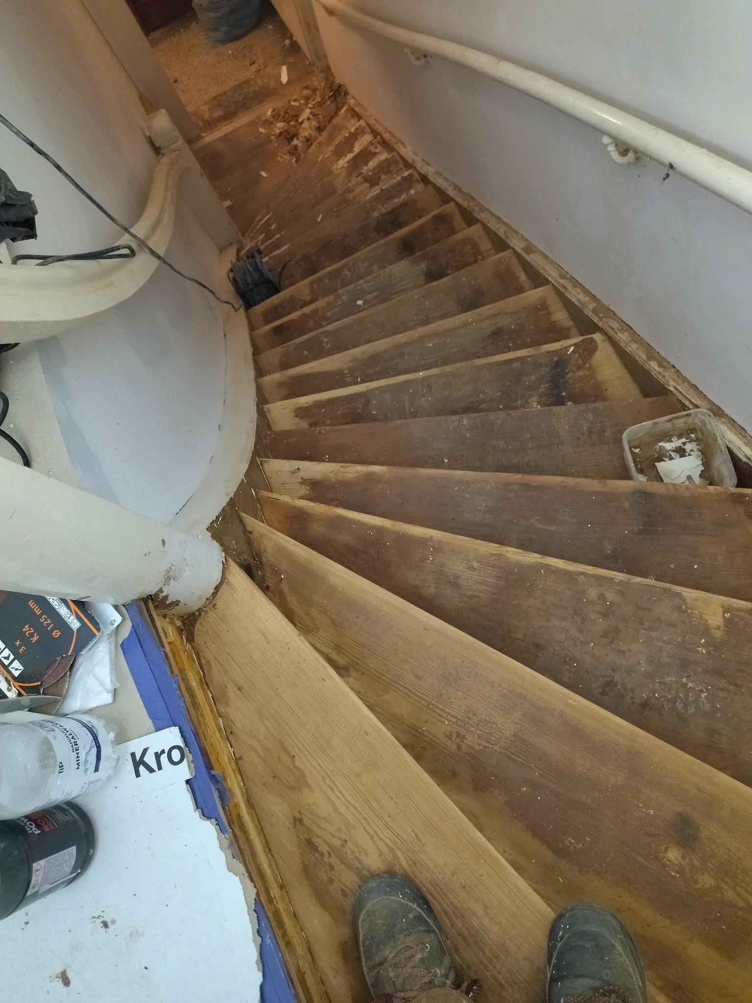 Demontaż starych drewnianych schodów: widoczne uszkodzone stopnie, ślady po kleju, resztki materiałów budowlanych, narzędzia i buty robocze na dole kadru, ujęcie z góry w trakcie renowacji.