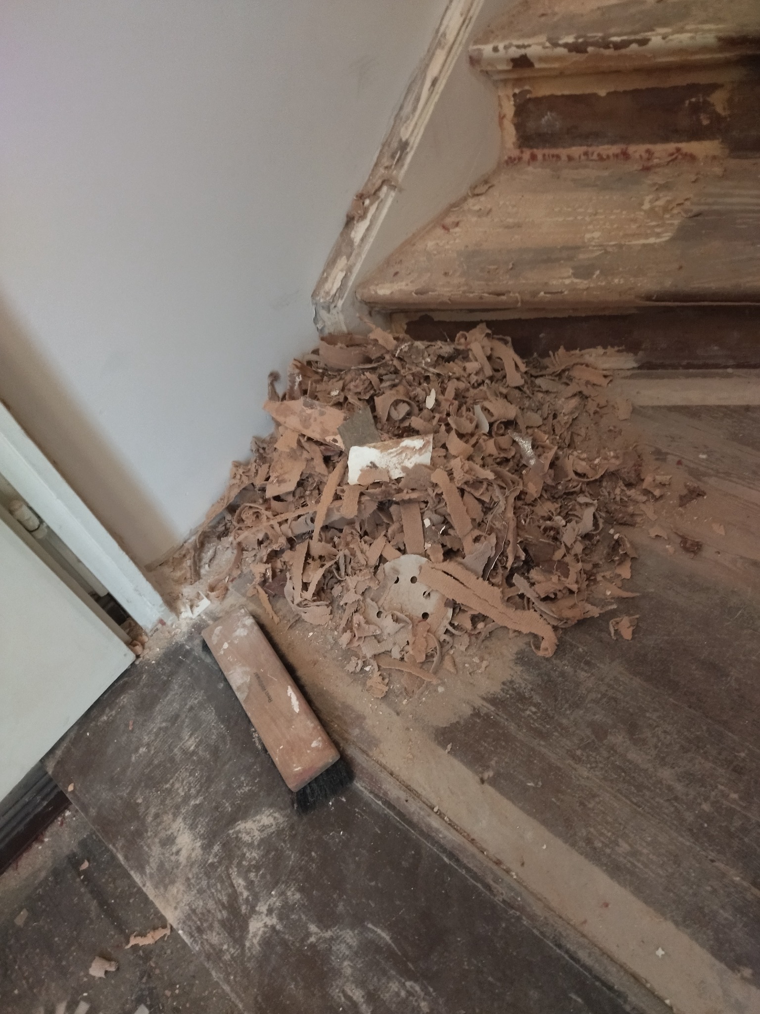 Fragment drewnianych schodów w trakcie renowacji, z kupką zeskrobanego starego lakieru i szczotką do zamiatania.