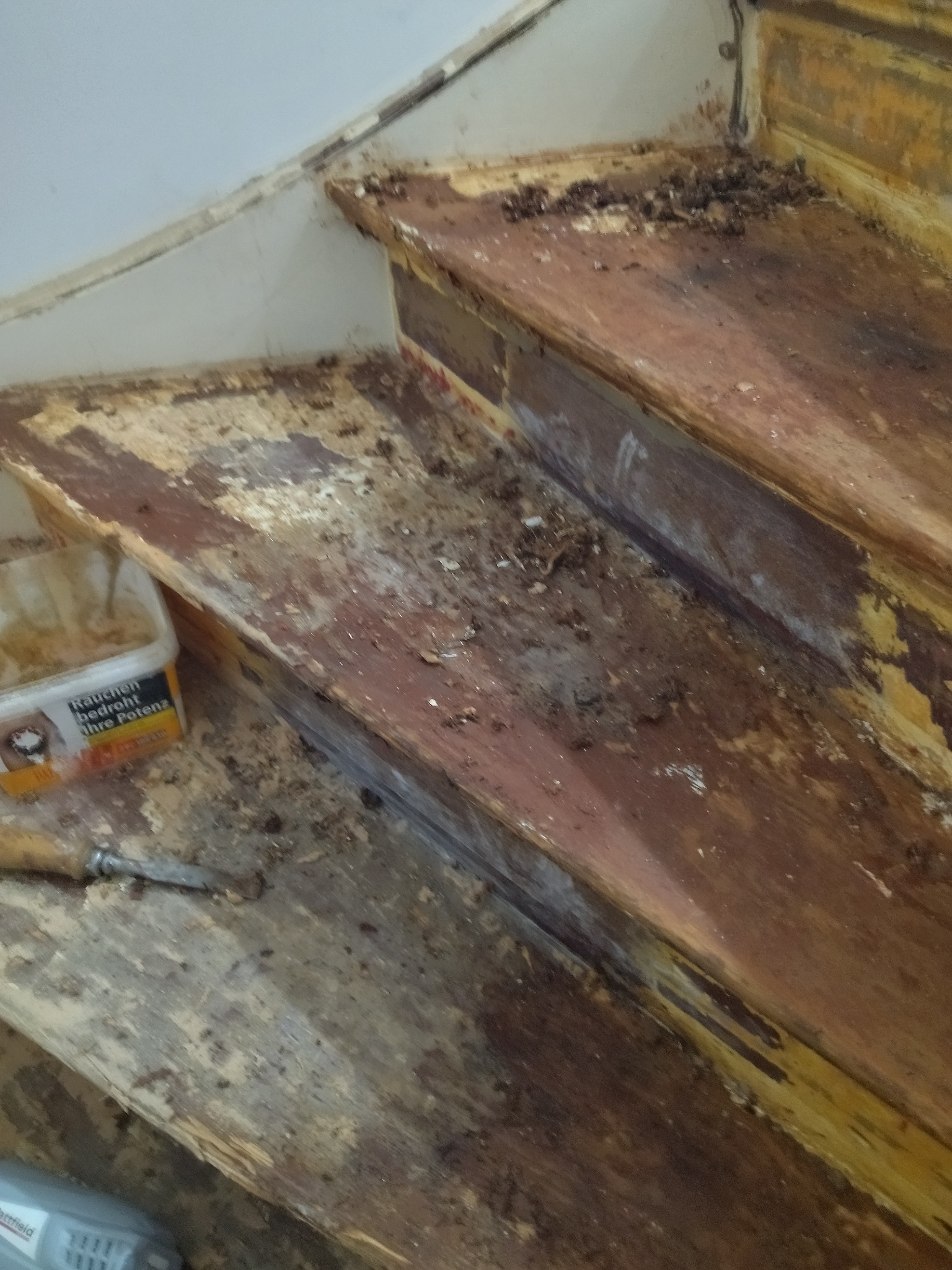 Demontaż starych drewnianych schodów: widoczne resztki kleju, odłupana farba, dłuto i pojemnik z rozpuszczalnikiem, przygotowanie do renowacji.