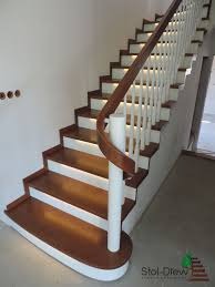 Bieg schodów z drewnianymi stopniami w kolorze orzecha, białymi podstopnicami i balustradą z białymi tralkami oraz drewnianym pochwytem, oświetlony od dołu, widoczne gniazdka elektryczne na ścianie.