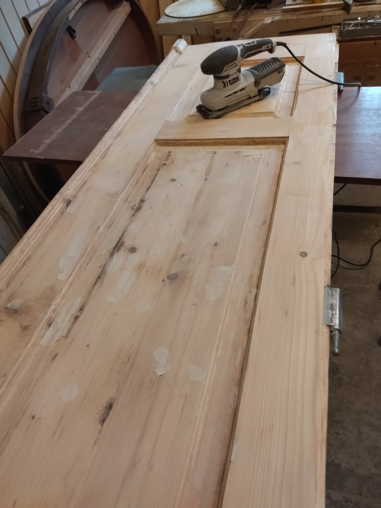 Szlifowanie drewnianych drzwi za pomocą szlifierki oscylacyjnej, widoczne ślady po szpachli, drzwi leżą na stole warsztatowym.