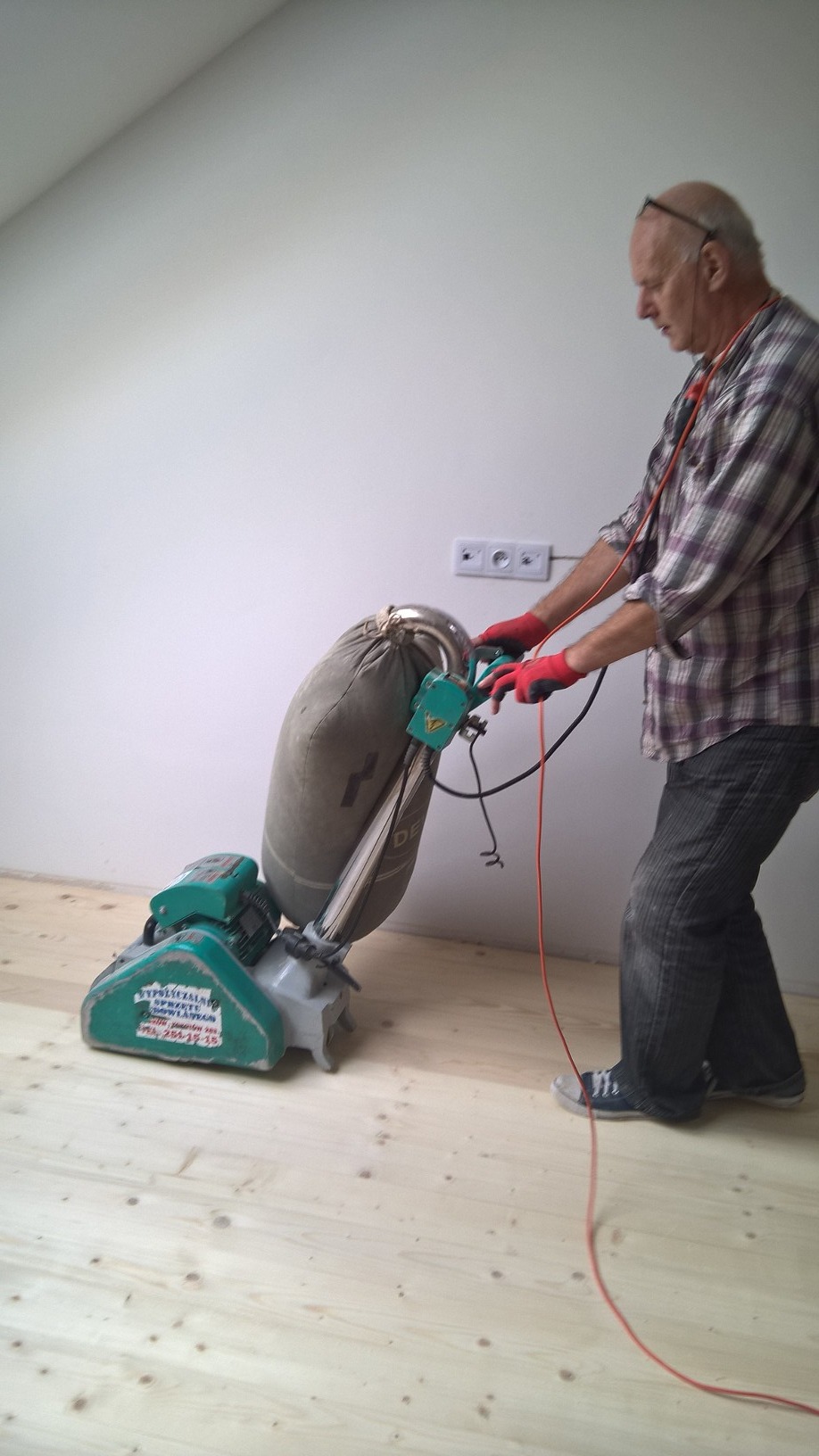 Mężczyzna w czerwonych rękawicach szlifuje drewnianą podłogę za pomocą profesjonalnej szlifierki podłogowej z workiem na pył, widoczne gniazdka elektryczne w tle.