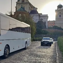 Biały autokar turystyczny na tle Starego Miasta w Warszawie, widok z zewnątrz i wnętrza z fotelami z logo firmy przewozowej.