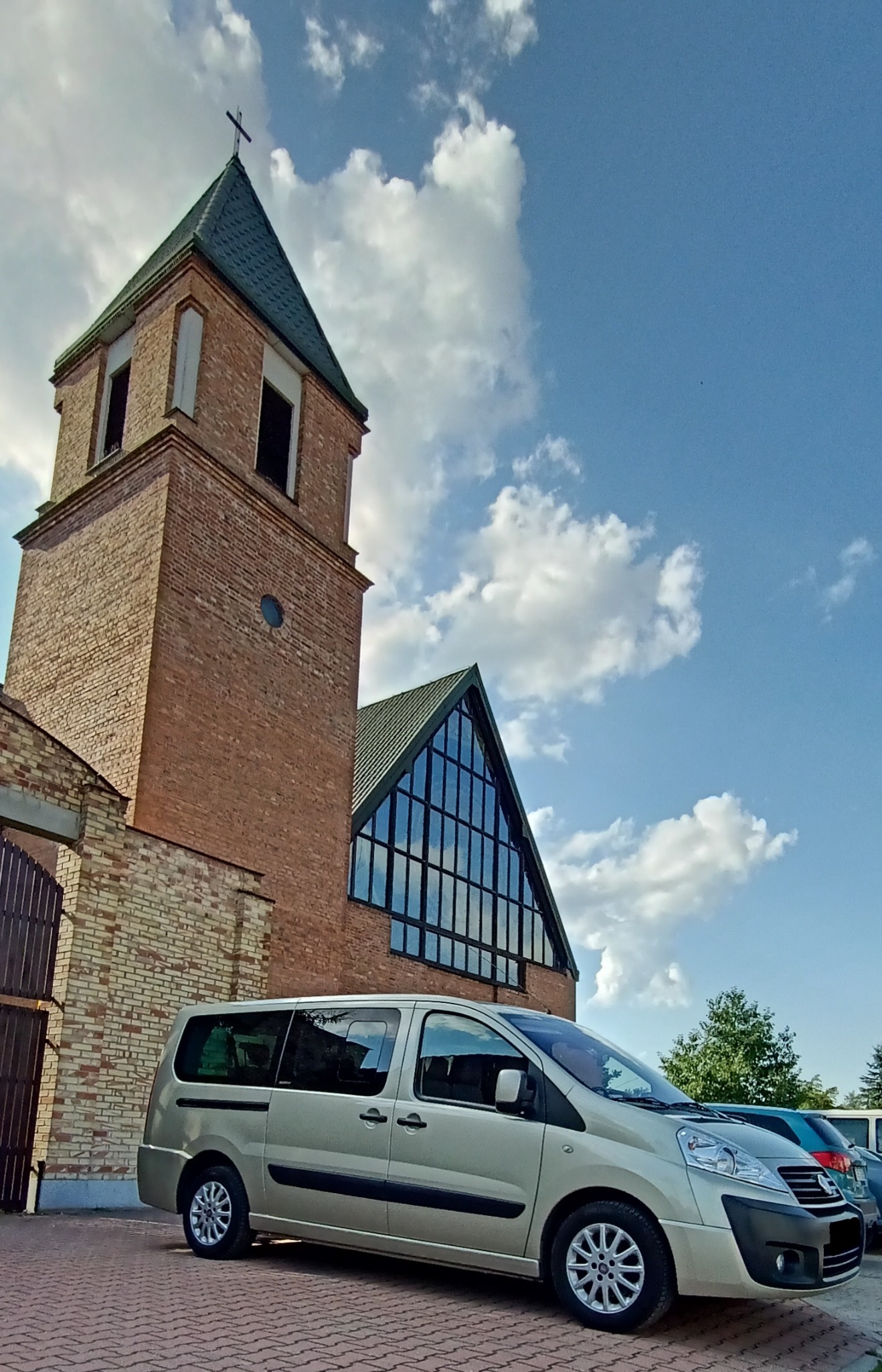 Beżowy van na brukowanym parkingu przed ceglanym budynkiem z wieżą zwieńczoną krzyżem, błękitne niebo z obłokami w tle.
