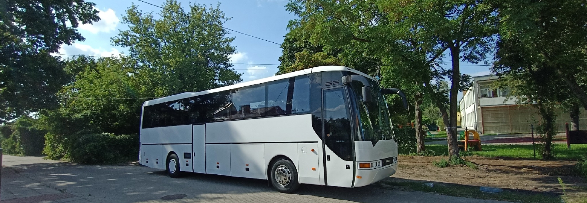 Biały autokar MAN zaparkowany na ulicy w słoneczny dzień, z zielonymi drzewami w tle i budynkiem w oddali.