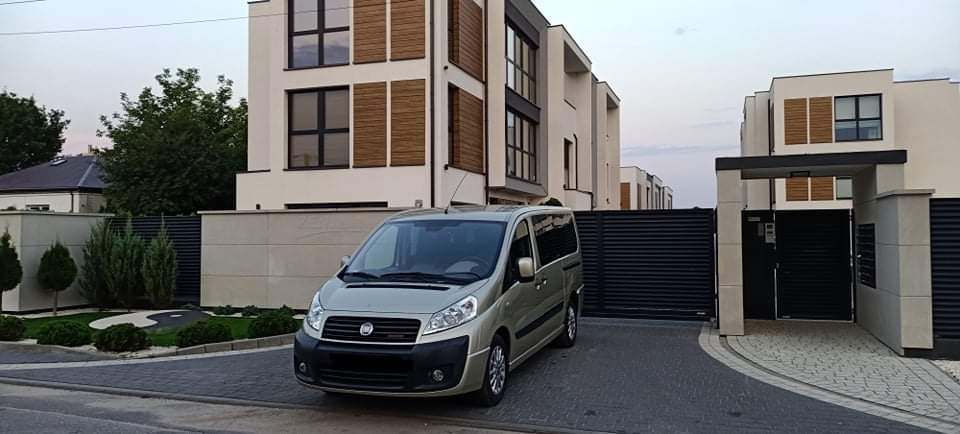 Szary van Fiat Scudo zaparkowany przed nowoczesnym domem z jasną elewacją i drewnianymi żaluzjami w oknach.