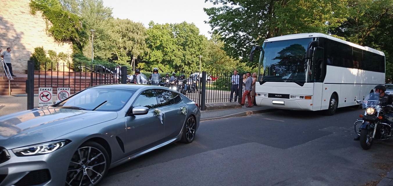 Srebrny samochód BMW z dekoracjami ślubnymi, biały autokar MAN, grupa motocyklistów i goście weselni przed budynkiem z zakazem wprowadzania psów.