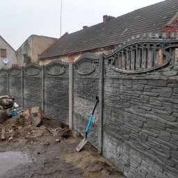 WIX-ART Brukarstwo - Nowo postawiony, betonowy płot z elementami dekoracyjnymi na tle zabudowań, z niebieską łopatą opartą o jeden z filarów i stertą gruzu obok.