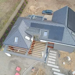 Widok z góry na dom z szarą dachówką, panelami słonecznymi i częściowo zadaszonym tarasem w trakcie budowy, widoczny samochód i kostka brukowa.