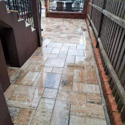 Świeżo ułożona, mokra kostka brukowa w odcieniach beżu i brązu, przylegająca do schodów z ciemnego kamienia i drewnianego ogrodzenia z elementami ceglanymi.