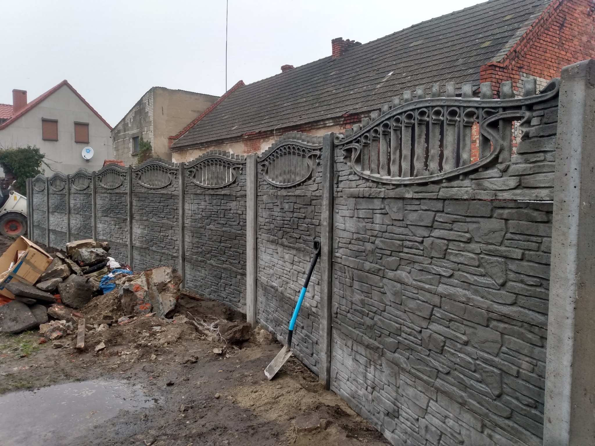 Nowo postawiony, betonowy płot z elementami dekoracyjnymi na tle zabudowań, z niebieską łopatą opartą o jeden z filarów i stertą gruzu obok.