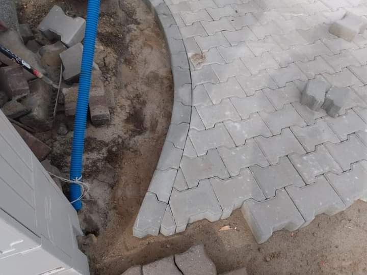 Fragment układanej nawierzchni z szarej kostki brukowej o nieregularnym kształcie, widoczna nieukończona krawędź, niebieska rura karbowana i piasek.