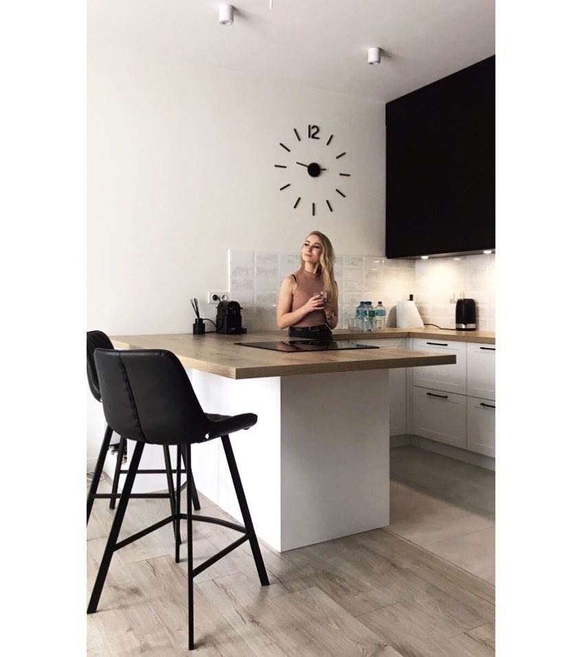 Nowoczesna kuchnia z wyspą, drewnianym blatem i czarnymi hokerami. Na ścianie duży zegar, a w tle kobieta trzymająca szklankę.