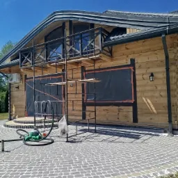 Drewniany dom z rusztowaniem, okna zabezpieczone folią, przygotowania do prac elewacyjnych, widoczny agregat do czyszczenia.