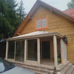 Drewniany dom z dobudowaną przeszkloną werandą w trakcie prac wykończeniowych, okna zaklejone folią ochronną, widoczna drabina i odkurzacz przemysłowy.