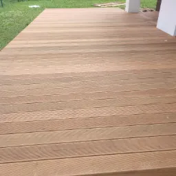 Nowo ułożony, jasnobrązowy taras z desek kompozytowych, widok z góry na całą powierzchnię, z fragmentem trawnika i elementami konstrukcyjnymi budynku w tle.