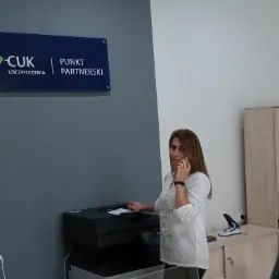 Pracownica punktu partnerskiego CUK Ubezpieczenia odbiera telefon przy drukarce, w tle ściana w dwóch odcieniach szarości.