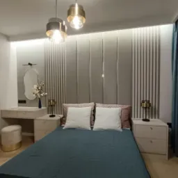 Nowoczesna sypialnia z dekoracyjnym panelem ściennym, toaletką i lampami wiszącymi. Stonowana kolorystyka, minimalistyczny design, przytulne oświetlenie.