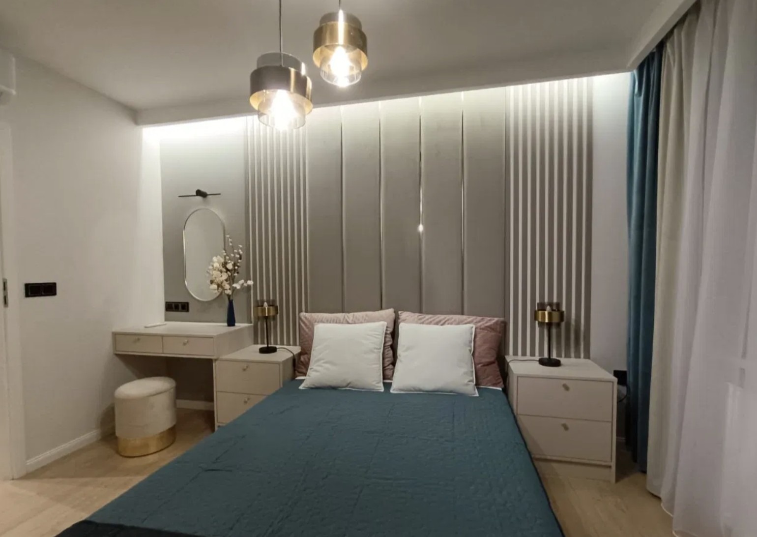 Nowoczesna sypialnia z dekoracyjnym panelem ściennym, toaletką i lampami wiszącymi. Stonowana kolorystyka, minimalistyczny design, przytulne oświetlenie.