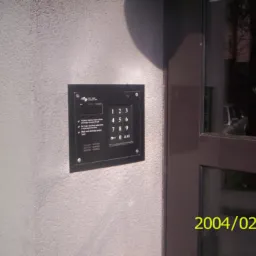 Czarny panel domofonu cyfrowego z klawiaturą numeryczną zamontowany na szarej ścianie obok brązowych drzwi wejściowych.