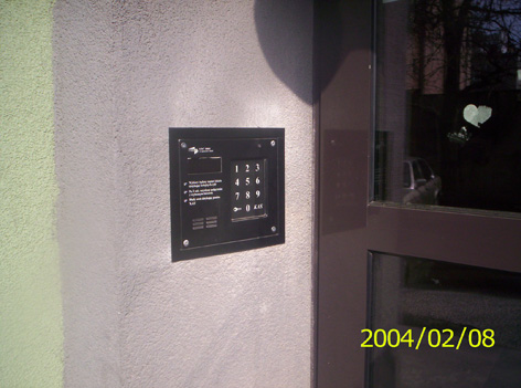 Czarny panel domofonu cyfrowego z klawiaturą numeryczną zamontowany na szarej ścianie obok brązowych drzwi wejściowych.