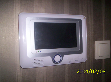 Zamontowany wideofon cyfrowy z białym obramowaniem na ścianie z tapetą w paski, obok włącznik światła. Data na zdjęciu: 2004/02/08.