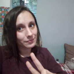 Kobieta z ciemnymi włosami, ubrana w bordową bluzkę, robi sobie selfie na tle szarej ściany z dekoracjami i poduszką, pokazując dwa palce.