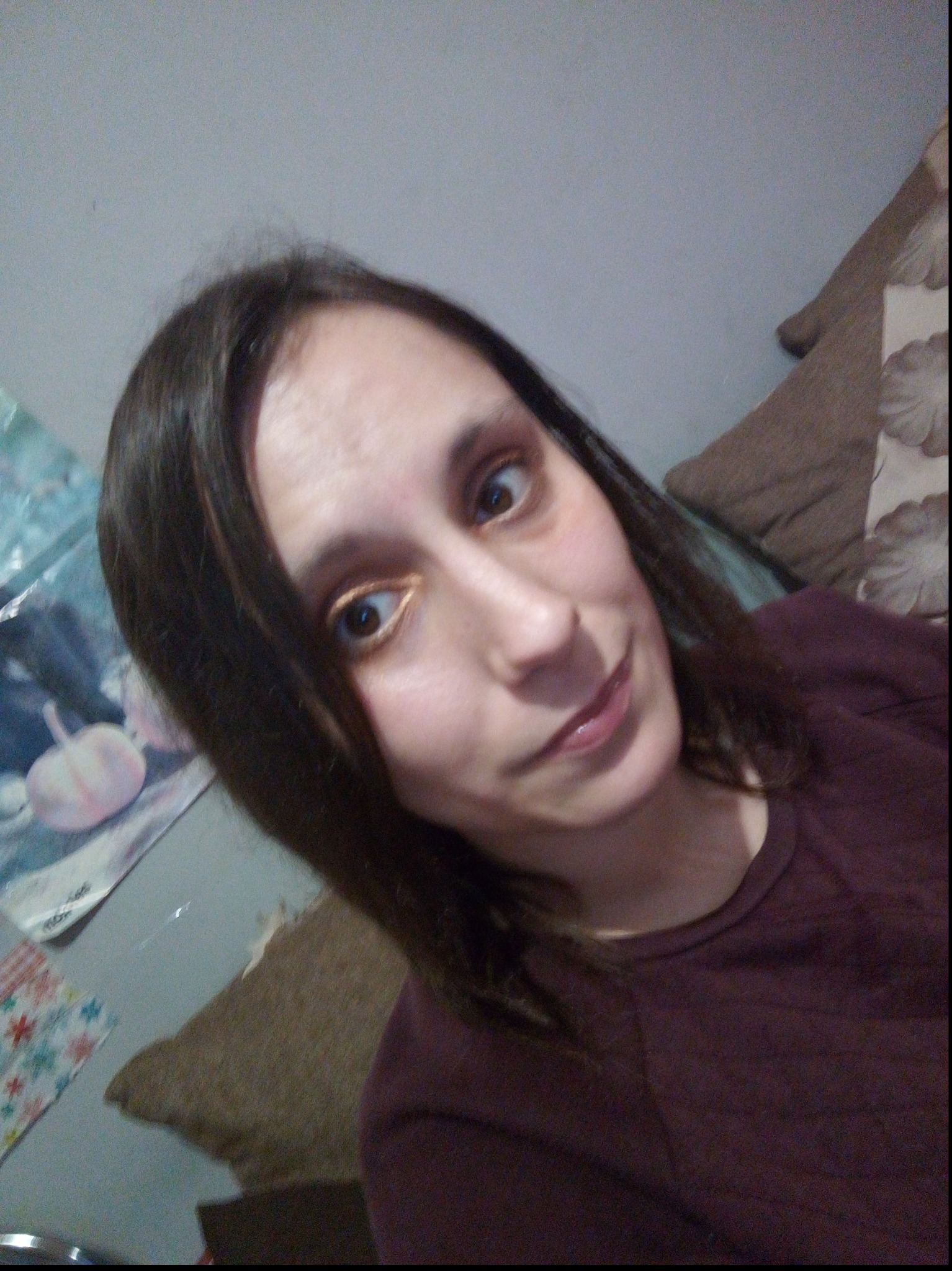 Portret kobiety z Sieradza, z lekkim uśmiechem i złotym makijażem oczu, na tle ściany z naklejkami i brązową poduszką.