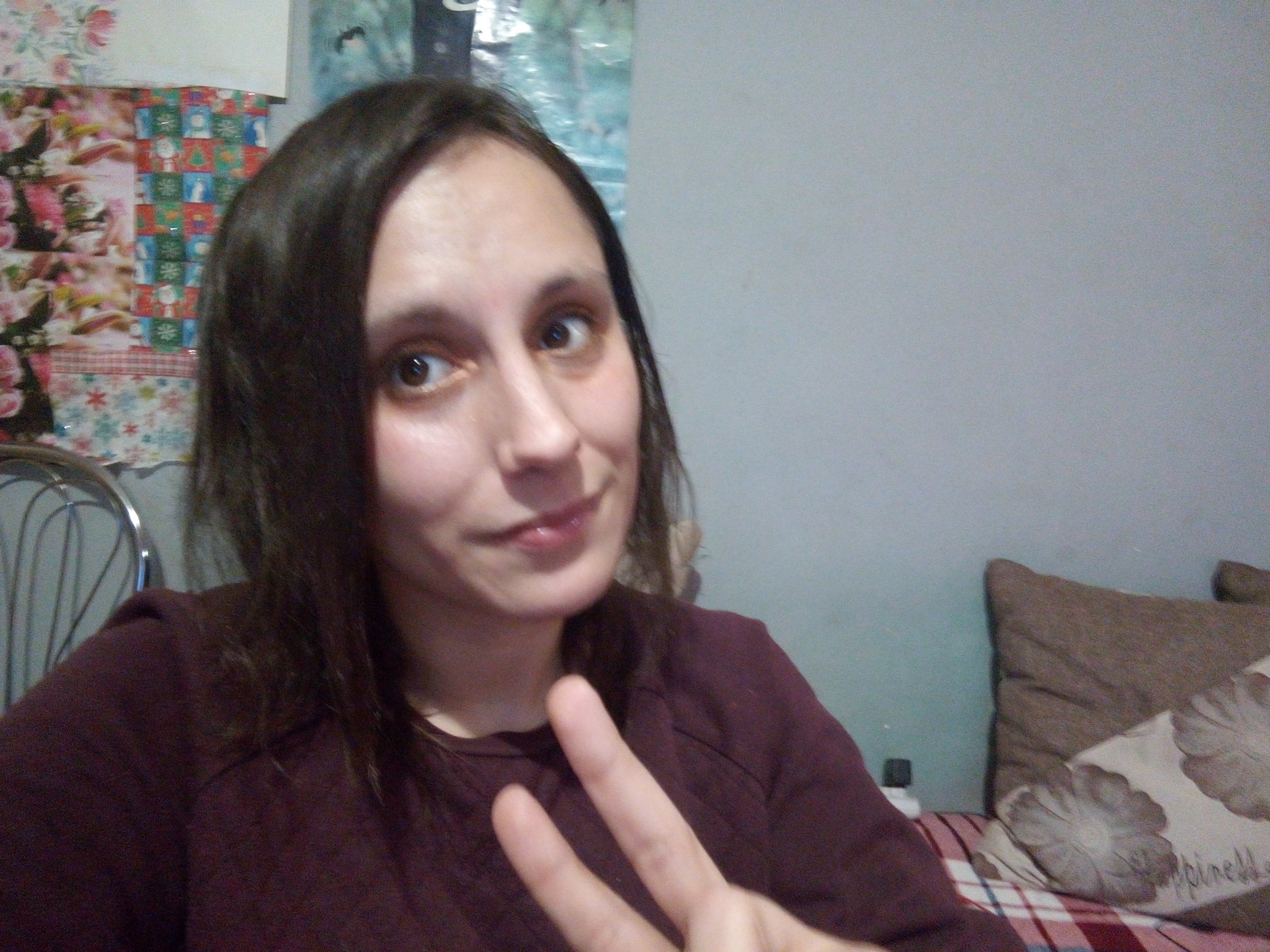 Kobieta z ciemnymi włosami, ubrana w bordową bluzkę, robi sobie selfie na tle szarej ściany z dekoracjami i poduszką, pokazując dwa palce.