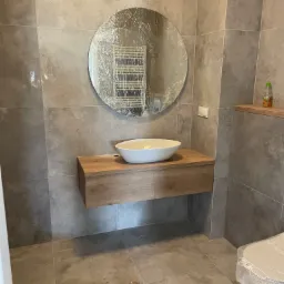 Nowoczesna łazienka z umywalką nablatową na drewnianej szafce, okrągłym lustrem w folii ochronnej i szarymi płytkami gresowymi na ścianach i podłodze. Ujęcie z widoczną toaletą również w folii...