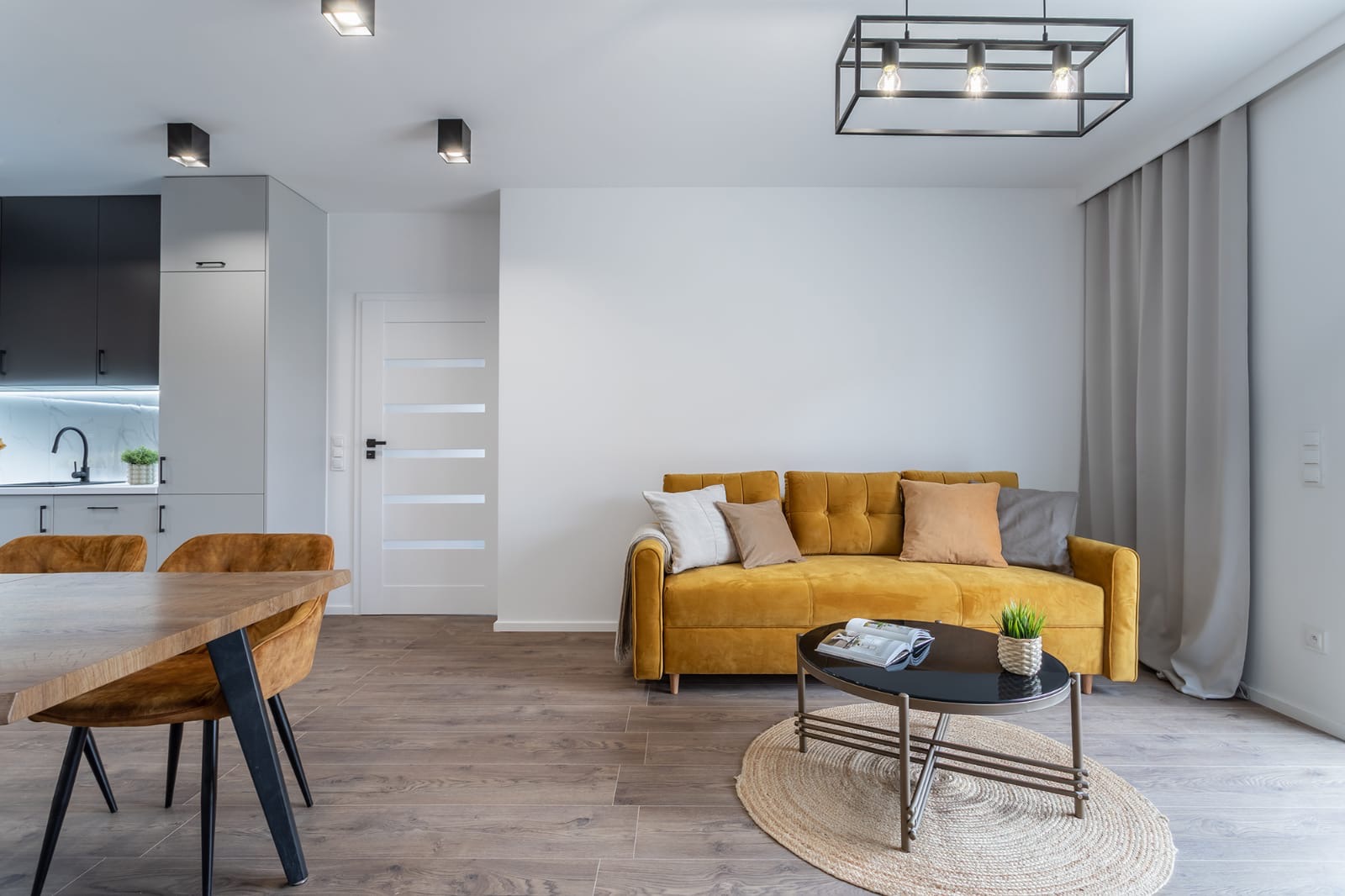 Jasne, nowoczesne wnętrze z drewnianą podłogą, musztardową sofą, okrągłym dywanem z juty, czarnym stolikiem kawowym, drewnianym stołem z krzesłami i szarą zabudową kuchenną.