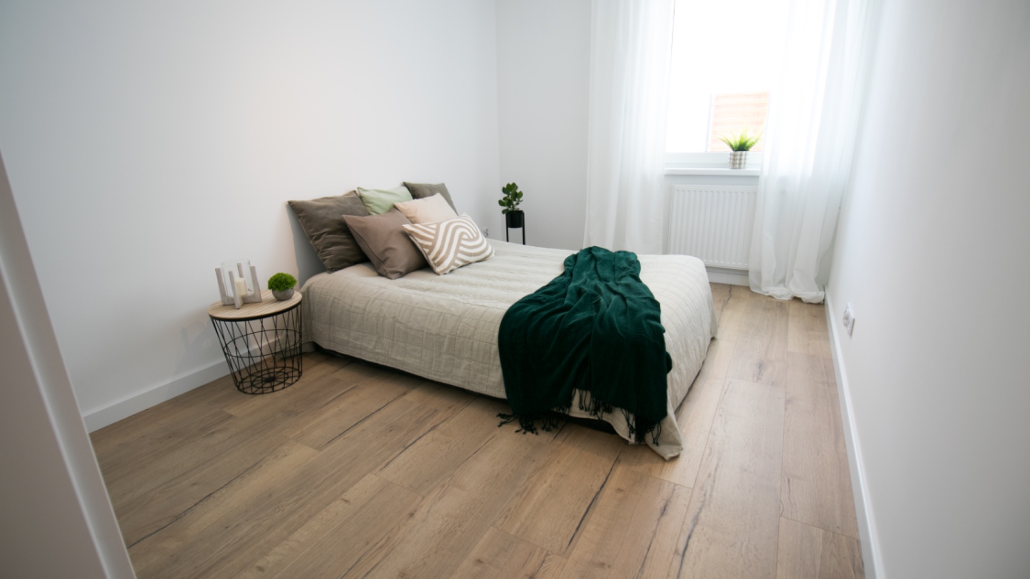 Jasna sypialnia z drewnianą podłogą, łóżkiem z zielonym pledem i stolikiem nocnym z dekoracjami, minimalistyczny wystrój.