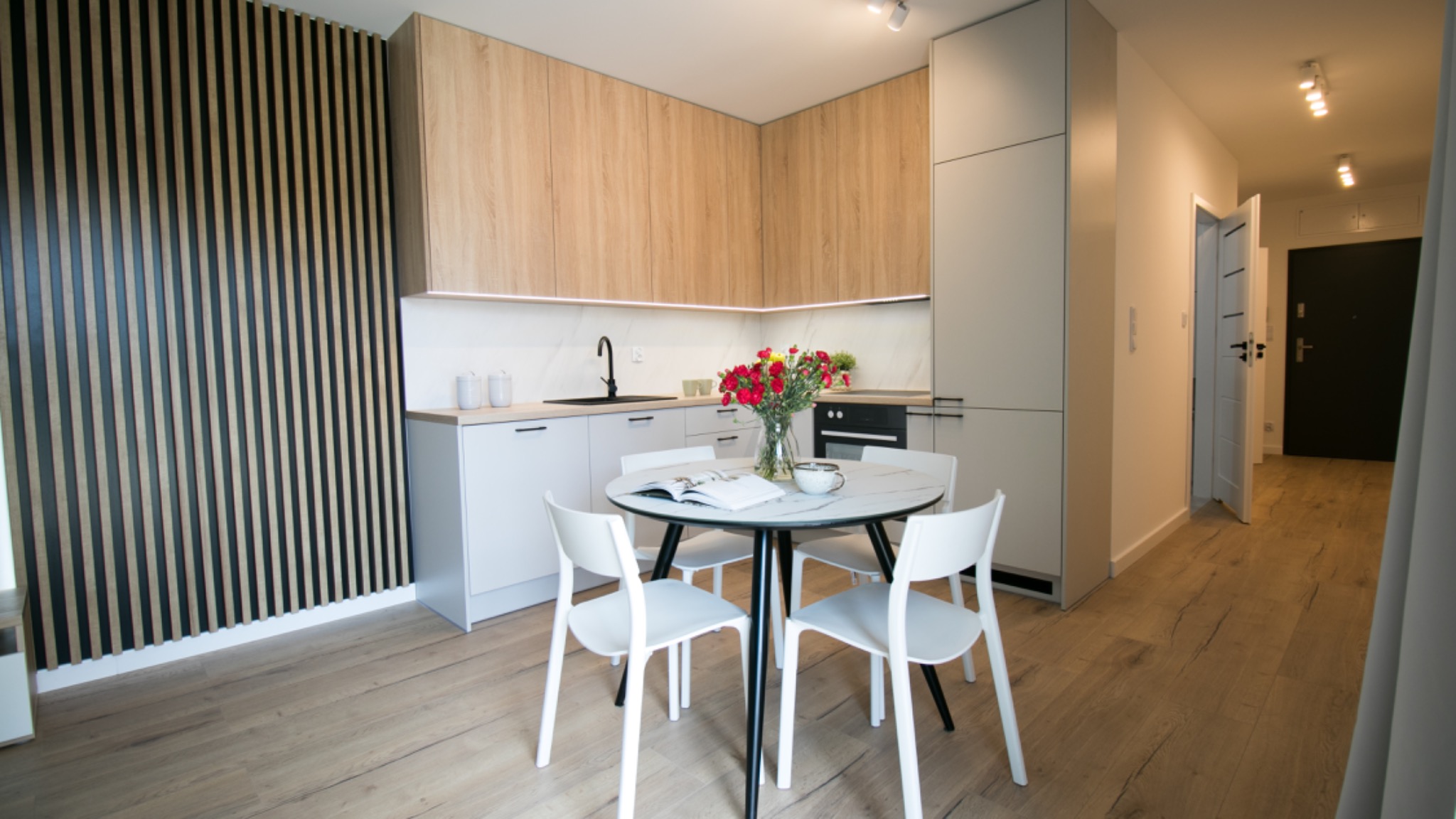 Jasne, nowocześnie urządzone mieszkanie z drewnianą podłogą, okrągłym stołem z marmurowym blatem i kwiatami, minimalistyczna kuchnia z drewnianymi szafkami i czarną armaturą, dekoracyjna ściana...