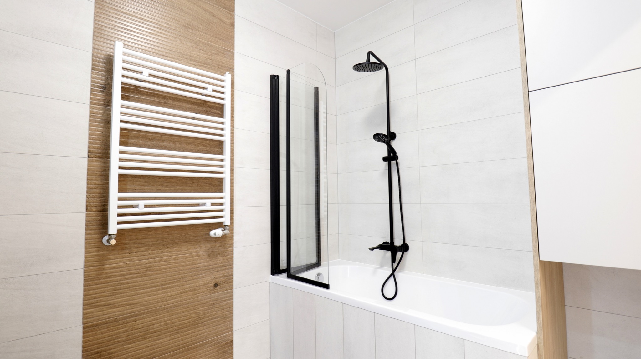 Nowoczesna łazienka z białą wanną, czarną armaturą, szklanym parawanem i grzejnikiem drabinkowym na ścianie wyłożonej płytkami imitującymi drewno.