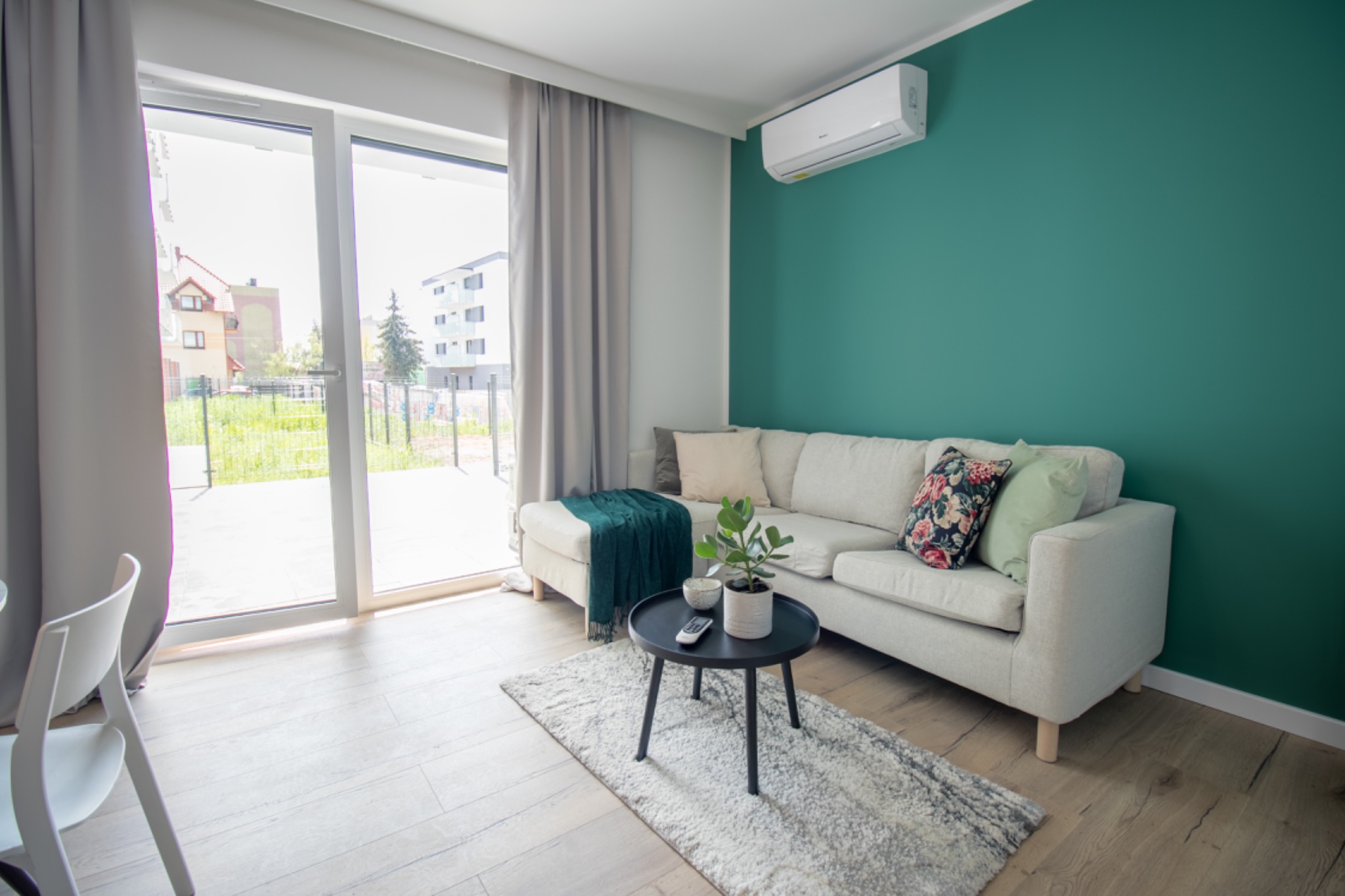 Jasne, nowocześnie urządzone wnętrze salonu z zieloną ścianą, beżową sofą i stolikiem kawowym, widok na zewnątrz przez przesuwne drzwi balkonowe.