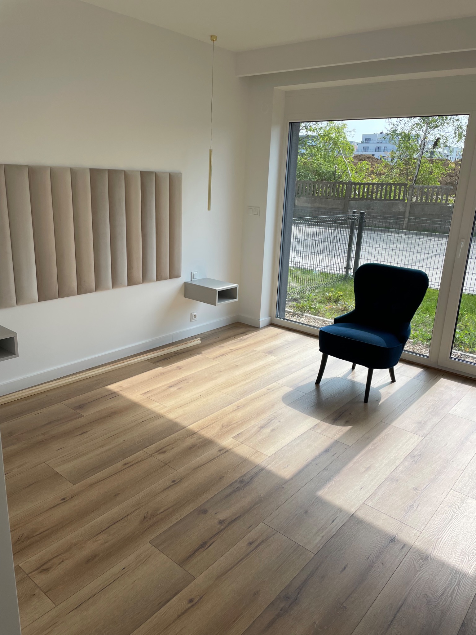 Jasne wnętrze pokoju z drewnianą podłogą, ciemnoniebieskim krzesłem obok okna, beżowym panelem ściennym i minimalistyczną lampą wiszącą