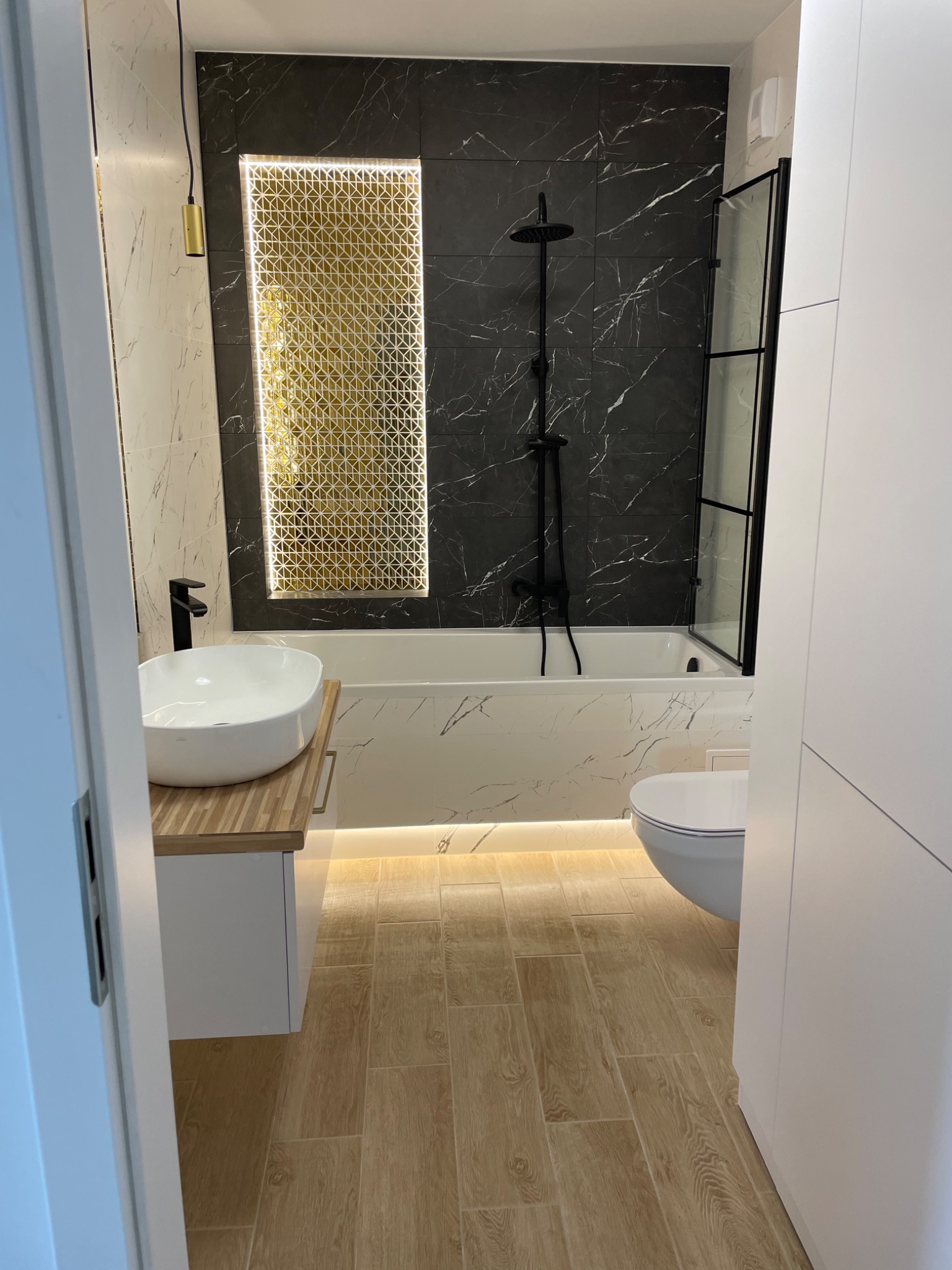 Nowoczesna łazienka z czarno-białymi marmurkowymi płytkami, złotym podświetlanym panelem dekoracyjnym, białą umywalką nablatową na drewnianym blacie i kabiną prysznicową z czarnymi ramami.