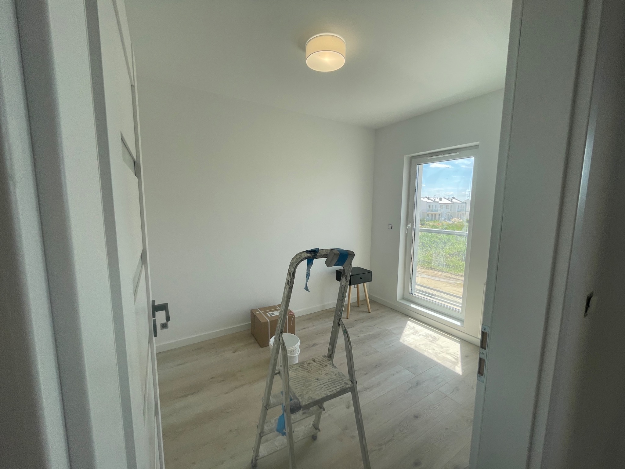 Pusty pokój z jasnymi ścianami, oświetleniem sufitowym i drabiną malarską na środku, przygotowany do malowania, z widokiem na osiedle domów przez okno.