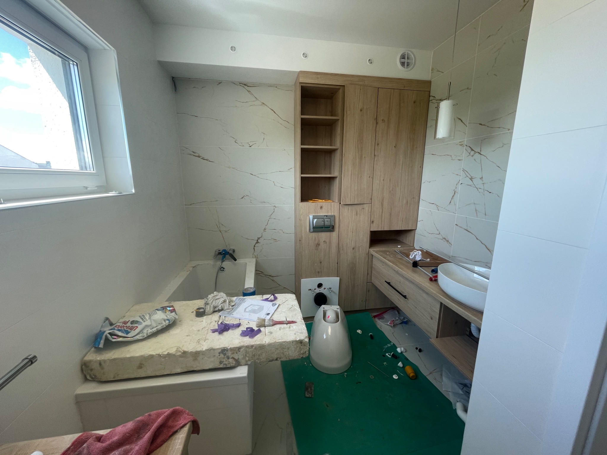 Łazienka w trakcie prac wykończeniowych: zamontowana umywalka, szafki z jasnego drewna, białe płytki z marmurkowym wzorem, niezabezpieczona toaleta i rozłożone narzędzia na zielonej folii ochronnej.