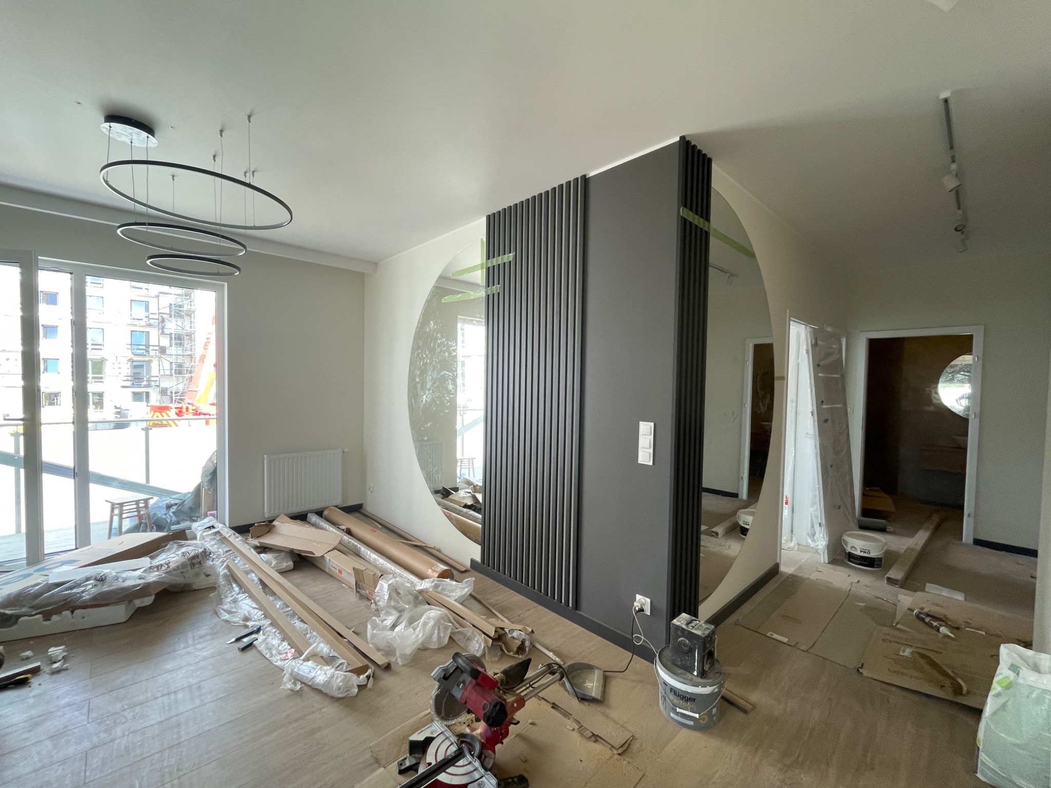 Wnętrze w trakcie prac wykończeniowych: widoczna szara ścianka działowa z pionowymi listwami, rozpakowane elementy wykończeniowe na podłodze, piła ukośnica i puszka farby Flugger.