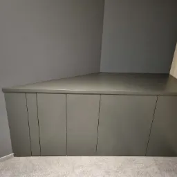 Szara, niska zabudowa meblowa w rogu pomieszczenia, z frontami bez uchwytów, dopasowana do skosu ściany, widoczne gniazdko elektryczne i fragment podłogi.