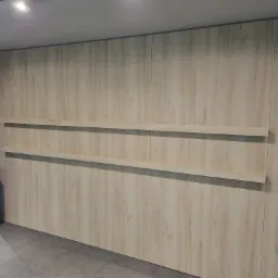 Ściana wykończona panelami drewnopodobnymi z dwoma półkami, oświetlenie sufitowe, fragment szafki z podświetleniem LED po lewej stronie.